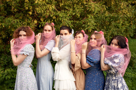 Charming bride in white dress with white veil and bridesmaids with pink veils lined up in a row, smiling and playfully covering their faces with veils. Cheerful bachelorette party in the summer park.の写真素材