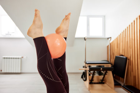 Cropped close-up of a womans legs holding an orange inflatable ball. A girl in a burgundy bodysuit does Pilates in a bright studio and trains muscle strength and coordination. Healthy lifestyle.の写真素材
