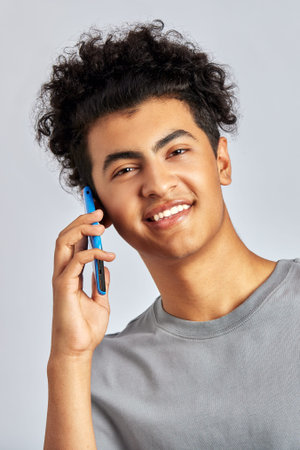 Studio portrait of a young handsome brunette man with curly hair holding a smartphone in his hand. A swarthy guy smiles and talks on a mobile phone. Modern lifestyle. Isolated on a gray background.の写真素材