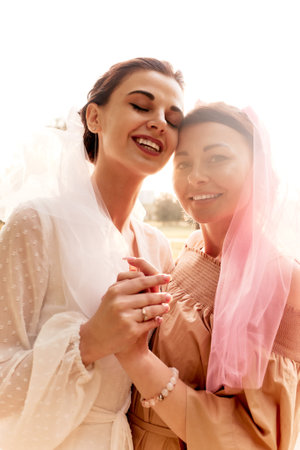Happy charming bride in a white dress with white veil and a bridesmaid in a beige dress with pink veil hugging tenderly and smiling. Cheerful bachelorette party. Close-up portrait.の写真素材