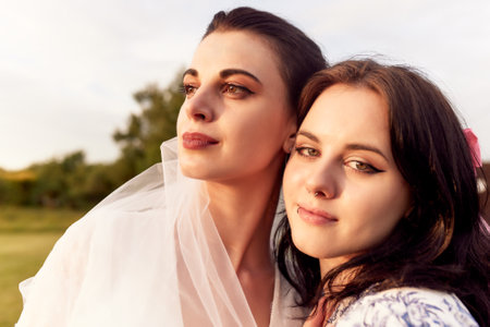 Close up portrait of happy charming bride in a white dress with white veil and a bridesmaid with pink veil hugging each other tenderly. Cheerful bachelorette party in a summer park.の写真素材