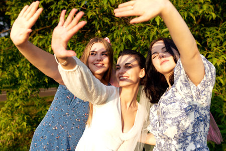 Charming young bride in a white dress and two bridesmaids with pink veils posing blocking their faces from the sun with their palms. Cheerful bachelorette party in the summer park. Close up portrait.の写真素材