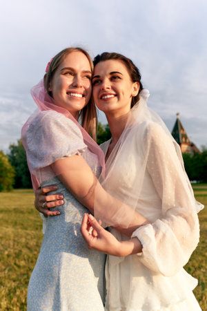 Half-length photo of charming bride in a white dress with a white veil and a bridesmaid in a light blue dress with pink veil gently hugging and smiling. Cheerful bachelorette party in the summer park.の写真素材