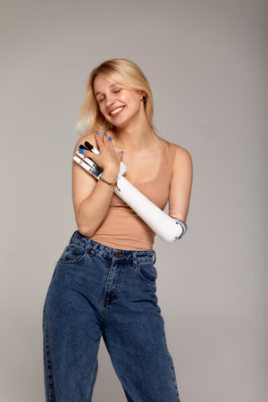 Studio half-length portrait of beautiful young blonde girl with disability wearing sensory bionic prosthetic arm. Woman dressed in blue jeans and beige top freely controls artificial robotic hand.の写真素材