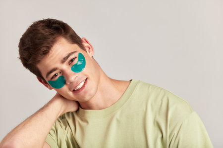 Young handsome smiling guy with brown hair applying under eye hydrogel patches on his face. Caucasian millennial man practices skin care routine to keep healthy and youthful looking. Studio portrait.の写真素材