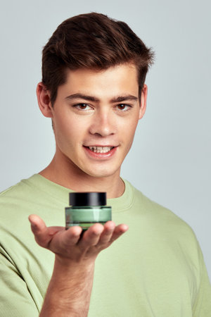 Young handsome guy with brown hair holds a small jar of moisturizing skin balm in his hand. Caucasian millennial man practices skincare routine to maintain healthy and youthful look. Studio portrait.の写真素材