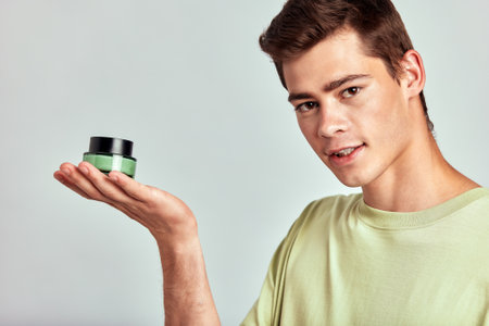 Young handsome guy with brown hair holds a small jar of moisturizing skin balm in his hand. Caucasian millennial man practices skincare routine to maintain healthy and youthful look. Studio portrait.の写真素材