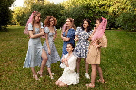 Happy charming bride in white dress with white veil sitting on the grass and bridesmaids with pink veils standing around her. Cheerful bachelorette party in a summer park.の写真素材