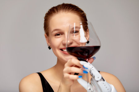 Studio portrait of a young girl with disability wearing sensory bionic prosthetic arm. Beautiful woman holds a glass of red wine with her artificial robotic hand. Full life after limb loss.の写真素材