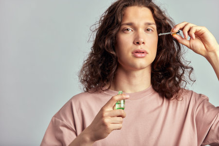 Young handsome long haired guy applying beauty serum on his face to keep his skin soft and smooth....