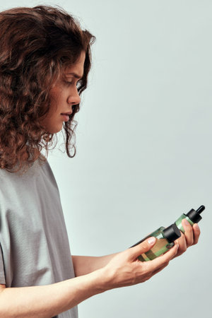 Portrait of young handsome long-haired guy looking at the bottles of moisturizing facial lotion and...