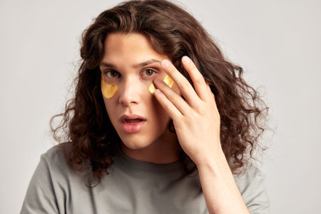 Young handsome long-haired guy applying under eye hydrogel patches on his face. Caucasian...