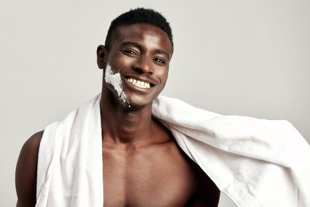 Studio portrait of handsome smiling black guy with naked torso, towel on his shoulder and cream on his face. Afro American man uses beauty products in his skincare routine to maintain healthy look.の写真素材