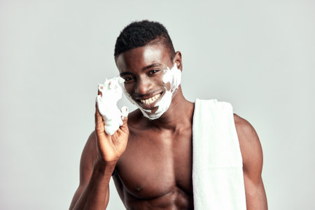 Studio portrait of handsome black guy with naked torso and towel on his shoulder. Happy smiling Afro American man applies cream on his face during his skincare routine to maintain his healthy look.の写真素材