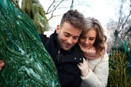 Beautiful young couple in love buying a Christmas tree from the market to decorate their home. Caucasian man and woman are preparing for Christmas. Anticipation of the New Year Eve in a snowy city.の写真素材