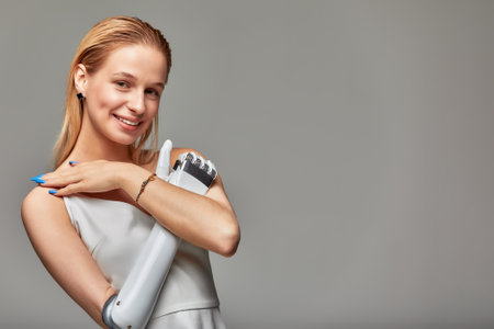 Studio portrait of beautiful young girl with disability wearing sensory bionic prosthetic arm. Smiling blonde woman in white dress crossed hands on her chest fully controlling artificial robotic hand.の写真素材