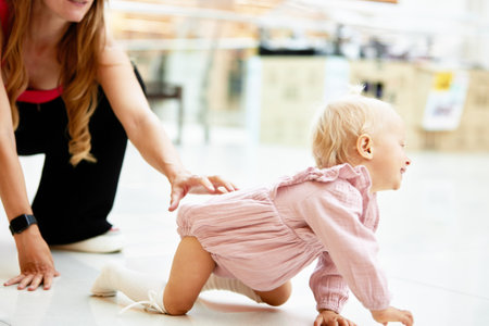 Happy young mother watch her cute baby crawling on floor at public place, shopping mall. Mom and her little daughter in pink clothes walking around mall and having fun. Funny weekend, happy childhood.の写真素材