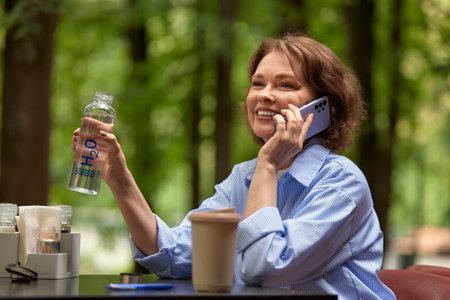Attractive mature woman sitting in cafe at the table with bottle of water and paper coffee cup....