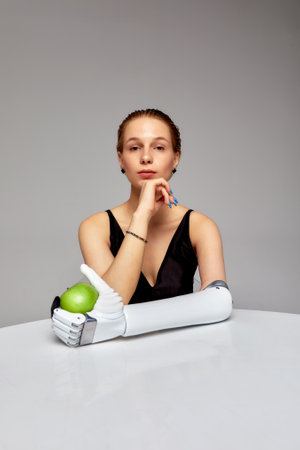 Young charming girl with disability wearing sensory bionic prosthetic arm seats at the table. Beautiful woman holds a green apple in her artificial robotic hand. Full life after limb loss.の写真素材