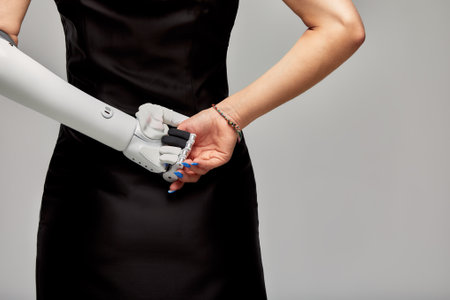 Cropped studio portrait of charming girl with disability wearing sensory bionic prosthetic arm. Slender woman in black dress stands her back to the camera with artificial robotic hand behind her back.の写真素材