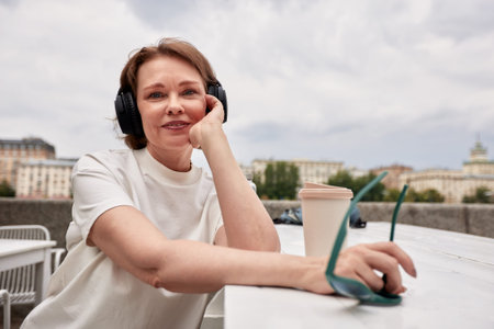 Adorable middle aged woman in white t-shirt, with sunglasses, wireless headphones and coffee paper...