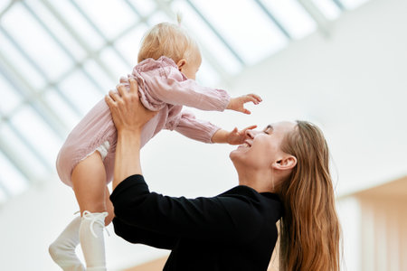 Young attractive mother with a baby in her raised hands. Happy mom holds her little daughter up in her arms. Tiny blond girl in pink clothes pretending to fly in mothers arms. Happy family.の写真素材