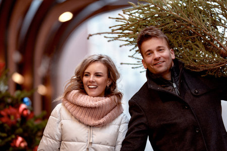 Beautiful happy young couple in love walking around Christmas market. Caucasian man and woman just bought Christmas tree and carry it home to decorate. Anticipation of the New Year Eve in snowy city.の写真素材