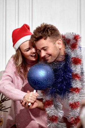 Beautiful romantic couple in love decorating Christmas tree at home. Happy man with garlands and woman in Santa hat preparing for Christmas and New Year celebration, having fun and fooling around.の写真素材