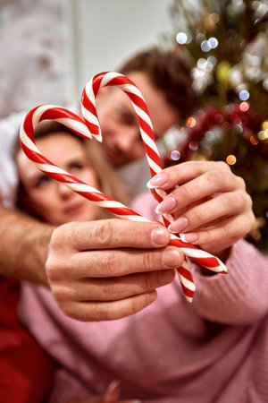Happy young couple in love lying in front of Christmas tree and hold candy canes in heart shape. Beautiful romantic couple preparing for Christmas and New Year celebration and having fun.の写真素材