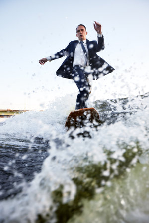 Young man in classic suit rides a wakeboard on the river or lake near city. A careless clerk escaped from a stuffy office to take up his favorite active sport. Best summer leisure after routine work.の写真素材