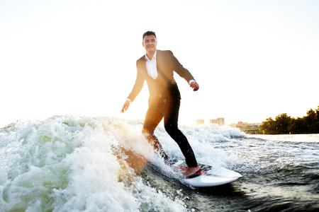 Young man in classic suit rides a wakeboard on the river or lake near city. Clerk escaped from a...