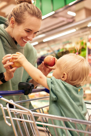 Happy young mother with her cute daughter in shopping trolley walking around supermarket. Beautiful mom and little blonde baby girl choosing fruit and having fun. Family shopping, happy childhood.の写真素材