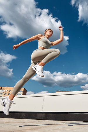 Fit woman exercising outdoors. Healthy young sportswoman doing fitness. An athlete makes jumps, does functional training on the street on a bright sunny day, smiles contentedly, in a sports suitの写真素材