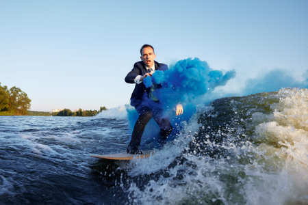 Young man in classic suit with blue smoke bomb in his hand rides wakeboard on a lake near city. Happy clerk escaped from stuffy office to take up active sport. Best summer leisure after routine work.の写真素材