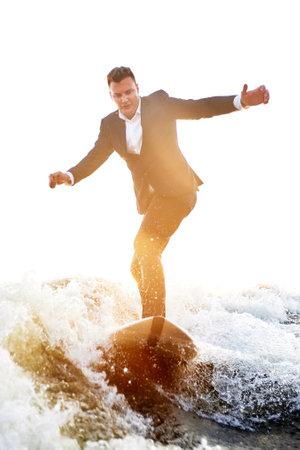 Young man surfer in classic suit rides on the wave. Happy clerk escaped from stuffy office to take up active sport. Best summer leisure after routine work. Film grain effect. Sun flare.の写真素材