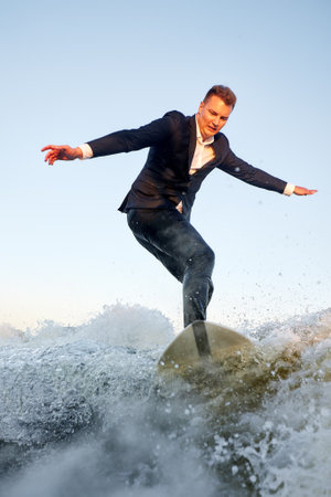 Young man surfer in classic suit rides on the wave. Happy clerk escaped from stuffy office to take up active sport. Best summer leisure after routine work. Film grain effect.の写真素材