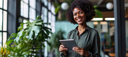 Half-length portrait of a beautiful young African American woman standing by the window in a bright modern startup office. Successful female professional uses tablet while working on a new project.の素材