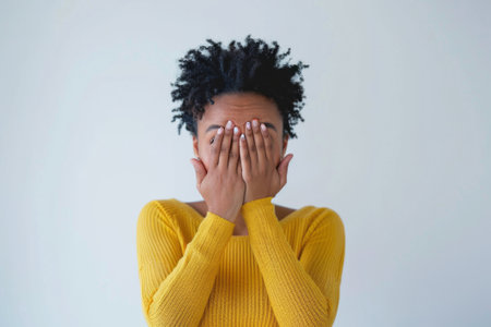 Studio portrait of attractive African American young woman in yellow blouse. Confused girl covered her face with her hands. Showing emotions. Isolated on white background.の素材