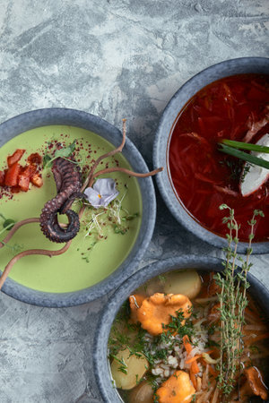 Gray plates with different soups on a gray background. Plate with asparagus soup with octopus, a plate of traditional borscht with sour cream, a plate with mushroom soup and standing on multi-colored linen napkins on a concrete marble gray background. Gray light texture background close-up top view. Beautiful table settingの写真素材