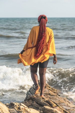 African woman walking on rocks by the beach in Accra. Has braided red rasta hair, Ghana West Africa.