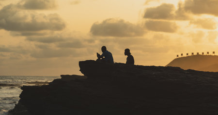 Sunset over Accra beach with rocks and two people sitting and enjoying the sea with hilly ground where you see the shooting range signs in the background. Accra Ghana 2020 November 9のeditorial素材