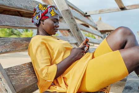 Ghana woman with african colorful headdress sitting on a bench in the village of keta with cellphoneの写真素材