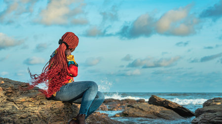 African woman with long red hair sitting on the beach rocks at sunset in Accra Ghana West Africaの写真素材