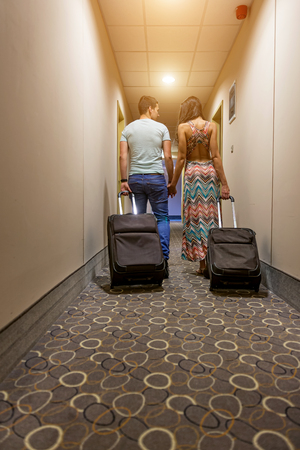 Young couple standing at hotel corridor upon arrival, looking for room, holding suitcases の写真素材