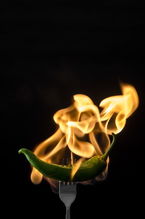 Green hot chili pepper on black background with flame on the forkの写真素材