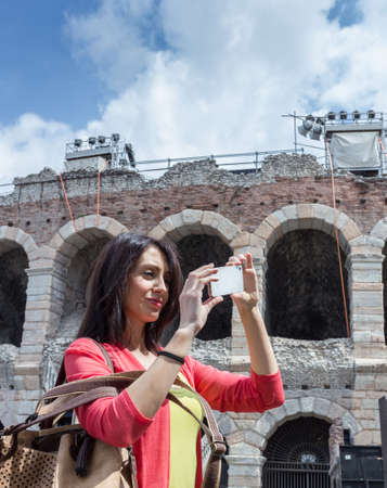 Female tourist taking a photo with mobile phone while sightseeing the city of Verona (Italy) with famous Arena on background. Shallow DOF.のeditorial素材