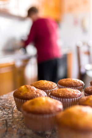 Batch of homemade freshly baked cupcakes or muffins cooling on a wire rack in the kitchen in a close up view with selective focus. Defocused blurry background. In the background the grandmother who has just cooked them.の写真素材