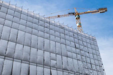 Building facade renovation with cellophane as protection and crane, against a blue sky with white clouds.の写真素材