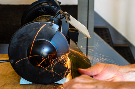 Man using a bench grinder to sharpen a lawnmower blade with sparks flying in many directions and...