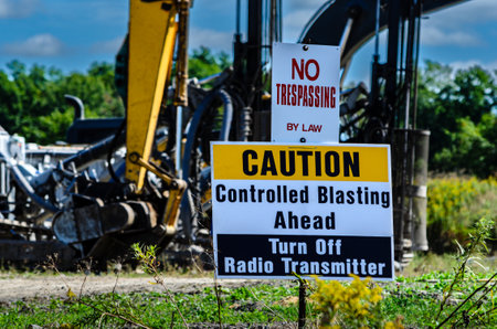 Close-up of a "Controlled Blasting" sign with Excavators and drilling machines in the background under a blue sky at a construction siteの写真素材